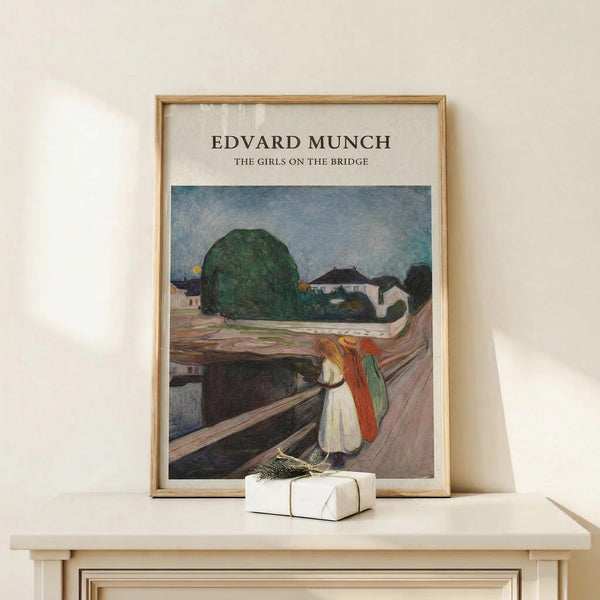 May include: Framed art print of Edvard Munch's expressionist painting showing three figures in colorful clothing standing on a wooden bridge, with green trees and white buildings in the background under a blue sky. The print has a cream mat and thin wooden frame, displayed on a light wood surface next to a small white wrapped gift with natural twine and greenery sprig.