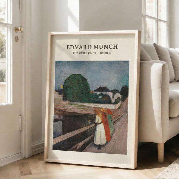 May include: Framed poster print of Edvard Munch's painting showing three girls in colorful dresses walking on a wooden bridge, with green trees and buildings in the background. The artwork features Munch's characteristic expressionist style with bold brushstrokes and muted colors. The print is displayed in a light wood frame, leaning against a white wall next to a beige sectional sofa in a bright, modern living room with large windows and natural lighting.