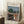 May include: Framed art print leaning against white wall corner showing Edvard Munch painting of three figures on wooden bridge with green tree and buildings in background. Light wooden frame, positioned on white textured rug near wooden side table with white coffee cup and saucer. Cream colored blanket visible at right edge.