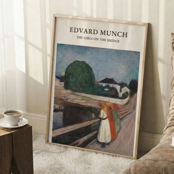 May include: Framed art print leaning against white wall corner showing Edvard Munch painting of three figures on wooden bridge with green tree and buildings in background. Light wooden frame, positioned on white textured rug near wooden side table with white coffee cup and saucer. Cream colored blanket visible at right edge.