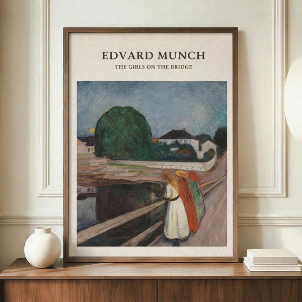 May include: Framed art print of Edvard Munch's "The Girls on the Bridge" displayed on wooden sideboard against cream paneled wall. Print shows three figures in white, red and green clothing standing on wooden bridge with large green tree and blue sky in background. Styled with white ceramic vase and stacked books. Warm wood frame complements modern interior setting.