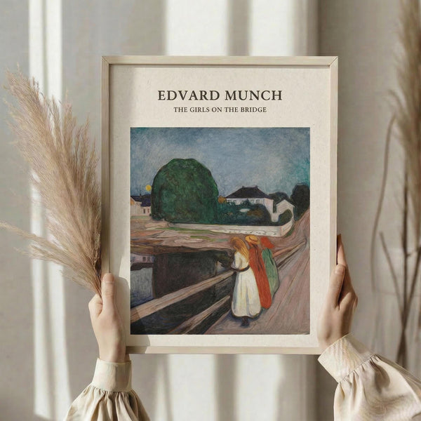 May include: Person in cream sweater holding framed art print featuring Edvard Munch's expressionist painting of figures on a wooden bridge, with houses and large green tree in background. Print has cream matting in light wood frame. Person also holds dried pampas grass stems against soft neutral curtained background.
