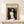 May include: Framed art print displaying Edvard Munch's Madonna artwork featuring a pale nude female figure with flowing dark hair against swirling background in black, white and orange tones, with small skull detail in corner. Print has cream matting in natural wood frame, positioned on light wooden surface beside small wrapped gift box against white wall.