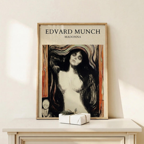 May include: Framed art print displaying Edvard Munch's Madonna artwork featuring a pale nude female figure with flowing dark hair against swirling background in black, white and orange tones, with small skull detail in corner. Print has cream matting in natural wood frame, positioned on light wooden surface beside small wrapped gift box against white wall.