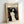 A framed art print leaning against a white wall shows Edvard Munch's expressionist lithograph featuring a nude female figure with long flowing dark hair against a swirling background. The artwork uses bold black lines and orange-red accents, with a small skeletal figure visible in the lower left corner. The print is displayed in a simple light wood frame on what appears to be a white surface.
