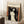May include: Framed art print featuring Edvard Munch's Madonna artwork showing a woman with flowing dark hair and pale skin against swirling black and orange background, with small skeletal figure in corner. Print has cream matting in light wood frame, leaning against white wall on textured white rug next to wooden side table with coffee cup and blanket.