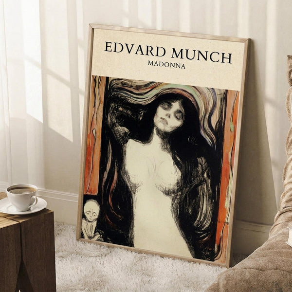 May include: Framed art print featuring Edvard Munch's Madonna artwork showing a woman with flowing dark hair and pale skin against swirling black and orange background, with small skeletal figure in corner. Print has cream matting in light wood frame, leaning against white wall on textured white rug next to wooden side table with coffee cup and blanket.