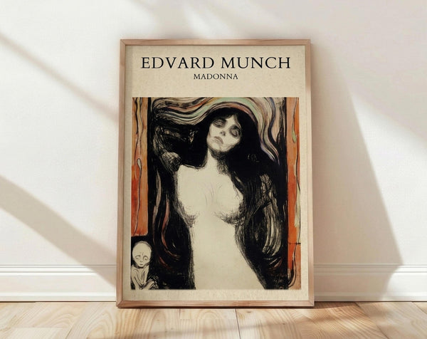 May include: Framed art print of Munch's Madonna lithograph featuring a pale nude woman with long flowing black hair against orange and red background, with small skeletal figure in corner. Print has cream colored mat in light wood frame, leaning against white wall on light wooden floor with soft natural lighting casting shadows.