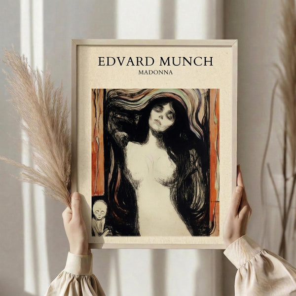 May include: Person in cream ruffled sleeves holding framed art print against white curtained window, with dried pampas grass visible. The poster features Edvard Munch's expressionist Madonna artwork showing a pale woman with flowing dark hair and closed eyes against orange and black swirling background, with small skeletal figure in corner. Artwork displayed in light wooden frame on cream mat.