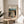 May include: Framed art print featuring Edvard Munch's expressionist painting of three figures in flowing white and yellow robes standing on a wooden bridge, displayed on a round wooden side table next to a beige upholstered chair with plant shadows cast on cream walls from natural sunlight.