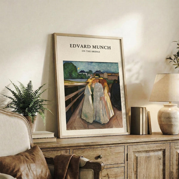 May include: Framed art print showing four figures in flowing white and yellow robes standing on wooden bridge planks against blue-green sky and distant buildings. Light wood frame with cream matting displayed on rustic wooden sideboard alongside fern plant, ceramic vase, stacked books, and beige upholstered furniture in neutral-toned living room setting.