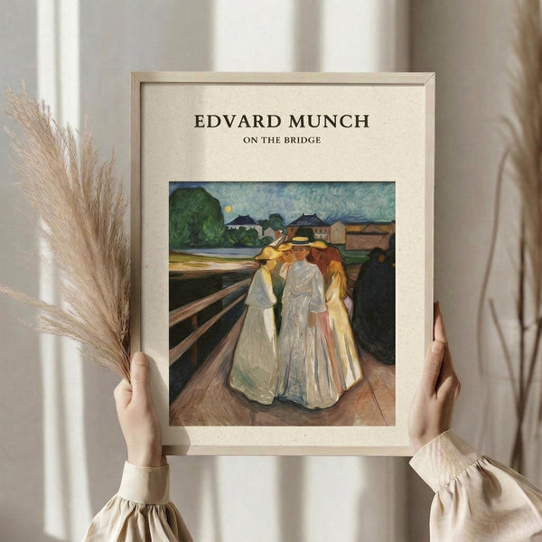 May include: Person in cream blouse with puffy sleeves holding framed art print and dried wheat stalks against white curtained window. The print shows Edvard Munch's painting of three figures in flowing white and yellow dresses standing on wooden bridge planks, with green landscape and buildings in background. Light wood frame with cream matting surrounds the artwork.