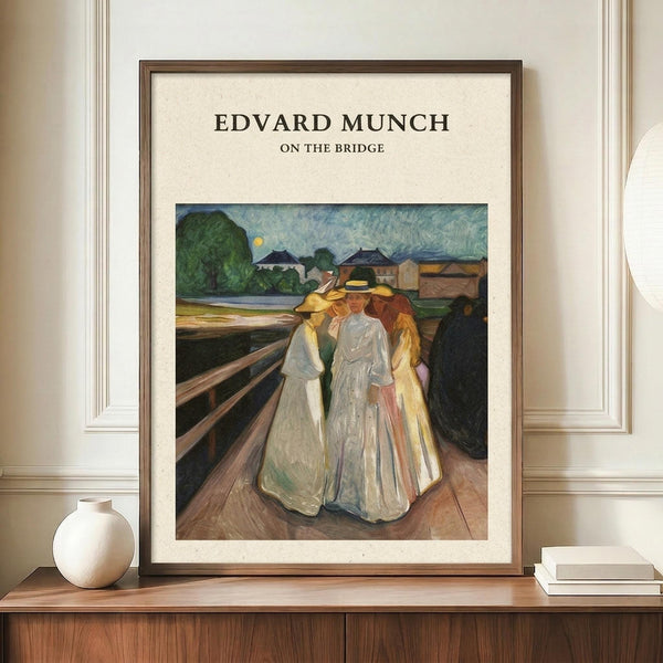 May include: Framed art print displaying Edvard Munch's painting of three figures in flowing white and yellow dresses standing close together on a wooden bridge, with green landscape and buildings in background under blue sky. Brown wooden frame with cream matting, displayed on walnut sideboard next to white ceramic vase and stacked books in neutral-toned room with white paneled walls.