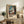 May include: Framed art print featuring a woman in white dress standing by blue window with curtains, painted in expressionist style with visible brushstrokes in blues, greens, and earth tones. Print displayed on rustic wooden console with books, fern plant, and ceramic vase in neutral-toned living room setting.