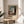 May include: Framed art print showing a young woman in white dress standing by a window with blue curtains and foliage. The expressionist painting features soft pastels and loose brushwork. The poster has museum-style text below the artwork. Displayed on a round wooden side table next to a beige upholstered chair with a throw pillow, palm plant visible, bright natural lighting creating shadows on white wall.