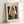 May include: Framed reproduction of Edvard Munch's "The Kiss" artwork in light wood frame, showing two nude figures embracing by a window with dark curtains, rendered in sepia and black tones with sketchy, expressive linework, displayed on white surface against white wall with soft natural lighting