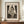 May include: Framed art print of Edvard Munch's "The Kiss" etching showing two embracing nude figures by a window with curtains, displayed on wooden sideboard next to white ceramic vase and stacked books in neutral-toned room with white paneled walls