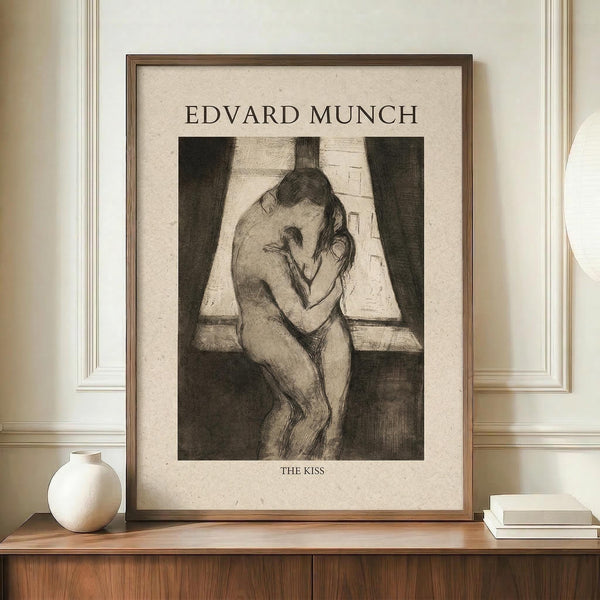 May include: Framed art print of Edvard Munch's "The Kiss" etching showing two embracing nude figures by a window with curtains, displayed on wooden sideboard next to white ceramic vase and stacked books in neutral-toned room with white paneled walls