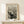 May include: Black and white poster print of Munch's The Scream artwork displayed in thin wooden frame on light wood surface against cream wall. Shows iconic figure with hands on face on bridge with swirling sky lines. Small wrapped white gift box with green sprig sits in front of frame.