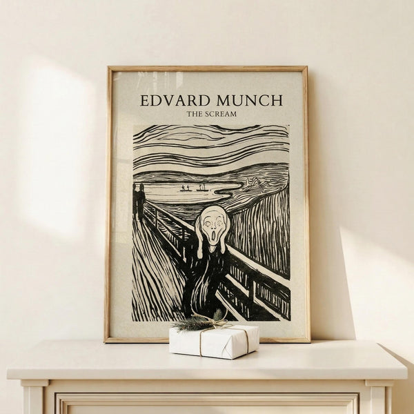 May include: Black and white poster print of Munch's The Scream artwork displayed in thin wooden frame on light wood surface against cream wall. Shows iconic figure with hands on face on bridge with swirling sky lines. Small wrapped white gift box with green sprig sits in front of frame.