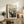 May include: Black and white print of The Scream artwork displayed in light wood frame on rustic wooden sideboard. Shows figure with hands to face on bridge with swirling sky lines. Room features cream chair with brown leather cushions, potted fern, ceramic vase, and stacked books against neutral walls.
