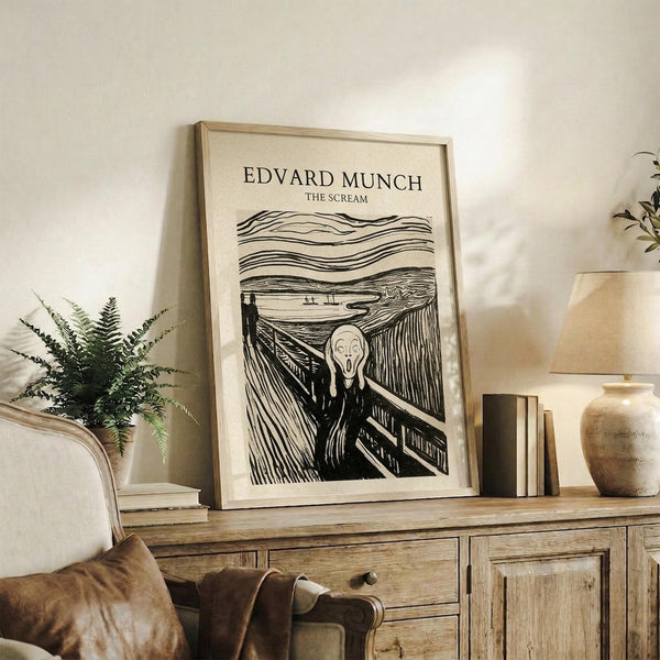 May include: Black and white print of The Scream artwork displayed in light wood frame on rustic wooden sideboard. Shows figure with hands to face on bridge with swirling sky lines. Room features cream chair with brown leather cushions, potted fern, ceramic vase, and stacked books against neutral walls.