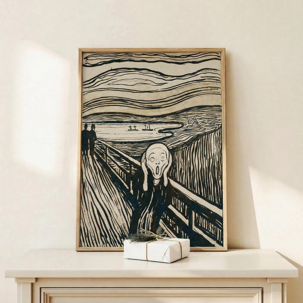 May include: Black and white reproduction of Edvard Munch's famous expressionist artwork displayed in thin wooden frame, leaning against cream colored wall on white wooden mantel or shelf. The iconic image shows a figure with skull-like face and open mouth on a bridge, with wavy lined sky and two distant figures in background. Small wrapped white gift box sits in front of the framed print.