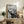 May include: Black and white line art reproduction of The Scream displayed in thin wooden frame on rustic wood sideboard. The expressionist artwork shows figure with hands to face on bridge with wavy sky lines. Room setting includes cream chair, fern plant, stacked books, ceramic vase, and neutral wall background.