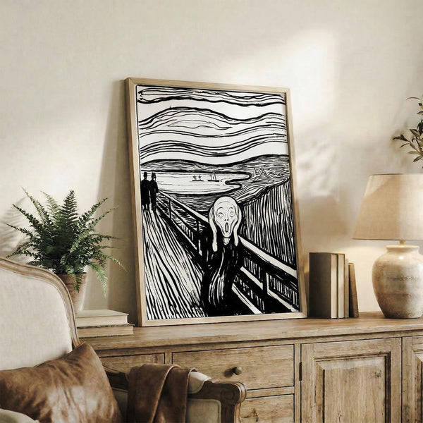 May include: Black and white line art reproduction of The Scream displayed in thin wooden frame on rustic wood sideboard. The expressionist artwork shows figure with hands to face on bridge with wavy sky lines. Room setting includes cream chair, fern plant, stacked books, ceramic vase, and neutral wall background.