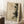 May include: Framed abstract art print leaning against white wall corner, featuring beige weathered background with dark green pine tree silhouette and geometric line elements including curved and straight marks. Natural wood frame, displayed on white textured rug beside wooden side table with white coffee cup, soft natural lighting creating window shadows on wall.