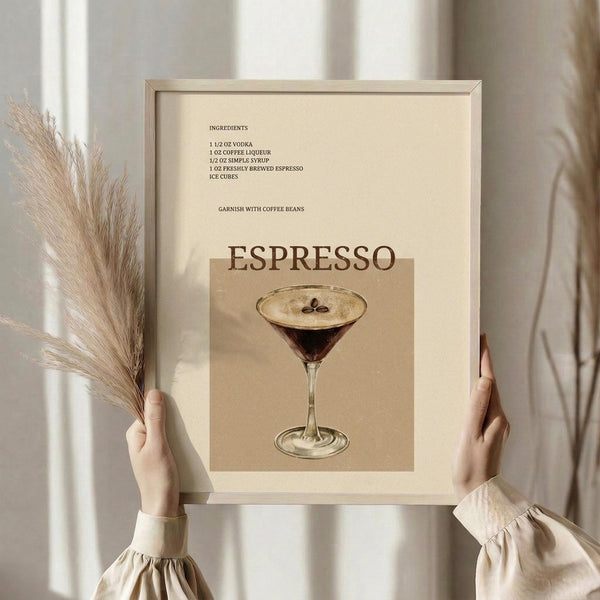 May include: Person in cream-colored top holding framed espresso martini cocktail recipe poster while holding dried pampas grass. The vintage-style print features a dark coffee cocktail in a martini glass with ingredient list on cream background. Light wooden frame against neutral wall with soft natural lighting.