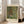 May include: Framed art print leaning against white wall next to beige sofa in bright living room. Print shows colorful garden scene with sunflowers, wildflowers, and lush greenery in impressionist style. White matted frame displays museum-style poster with cream background and black text identifying the artwork and artist Gustav Klimt.