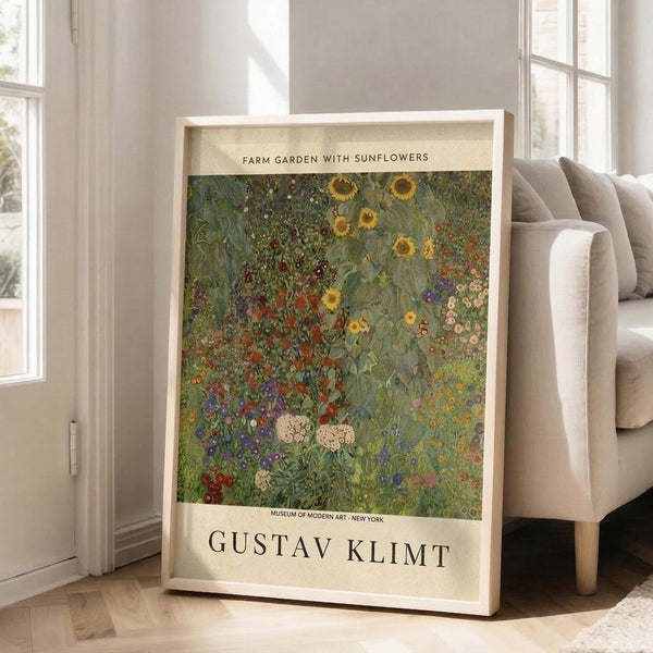 May include: Framed art print leaning against white wall next to beige sofa in bright living room. Print shows colorful garden scene with sunflowers, wildflowers, and lush greenery in impressionist style. White matted frame displays museum-style poster with cream background and black text identifying the artwork and artist Gustav Klimt.