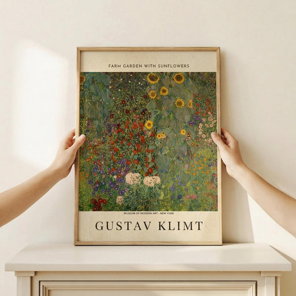 May include: Two hands holding a framed art print showing Gustav Klimt's colorful garden painting with vibrant sunflowers, wildflowers, and lush greenery in his distinctive decorative style. The print has museum-style text layout with cream matting and natural wood frame, displayed on a white wooden surface against a light wall.