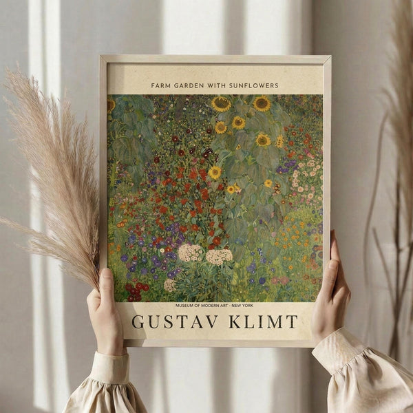 May include: Person holding framed art print featuring Gustav Klimt's colorful garden painting with sunflowers, wildflowers, and lush greenery in vibrant yellows, reds, purples, and greens. Light wood frame with cream matting displays museum attribution text. Dried wheat stems visible alongside, soft neutral clothing and curtained window background create warm, natural styling.