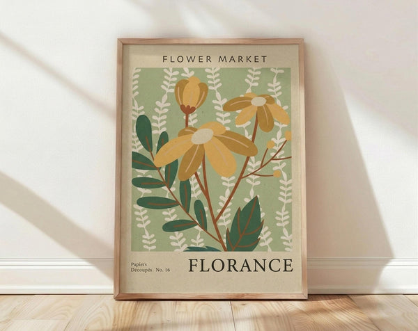 May include: Framed vintage-style botanical poster with sage green background featuring yellow daisy-like flowers and dark green foliage. Text reads "FLOWER MARKET" and "FLORANCE" in serif font. Light wood frame sits on wooden floor against white wall with soft natural lighting creating shadows.