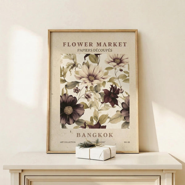 May include: Framed botanical art print featuring vintage-style illustrations of purple and cream flowers with green leaves on cream background. Displayed on light wood surface against white wall with small wrapped gift box nearby. Natural wood frame, classic exhibition poster design with Bangkok text.