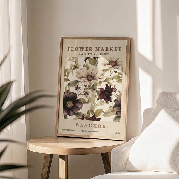 May include: Framed botanical poster with cream background displaying vintage-style illustrations of flowers in purple, white, and green tones. Text reads "Flower Market" and "Bangkok" in brown lettering. Wooden frame sits on round wooden side table next to beige upholstered chair. Natural sunlight streams across neutral-toned room with plant leaves visible in corner.