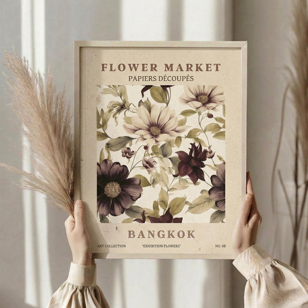 May include: Hands holding a framed botanical poster featuring vintage-style illustrations of white and deep purple flowers with green foliage on cream background. Text reads "Flower Market Bangkok" and "Papiers Découpés". Light wood frame, dried pampas grass visible to left, soft natural lighting against neutral curtained window backdrop.