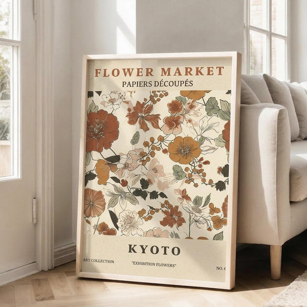 May include: Framed vintage-style floral poster with cream background featuring brown, orange, and green botanical illustrations. Text reads "Flower Market," "Papiers Découpés," and "Kyoto." Light wood frame leans against white wall next to beige upholstered chair in bright, airy room with large windows and hardwood floors.