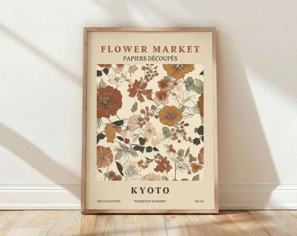 May include: Framed botanical art print with cream background featuring illustrated flowers in earthy tones of rust, orange, sage green, and brown. Text reads "Flower Market" at top and "Kyoto" at bottom. Displayed in thin wooden frame leaning against white wall on light wood flooring with natural shadows.