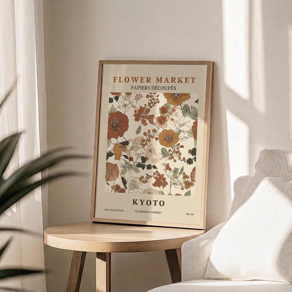 May include: Framed botanical art print featuring vintage-style flowers in warm autumn colors - rust, brown, gold, and green tones arranged in a floral pattern. The print has "FLOWER MARKET" and "KYOTO" text with decorative botanical illustrations. Wooden frame sits on a round natural wood side table next to a beige upholstered chair, with soft natural lighting and plant leaves visible in the corner of a neutral-toned room.