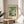May include: Botanical poster with white flowers and green foliage displayed in thin wooden frame on round wooden side table next to beige upholstered chair. Soft natural lighting creates window shadows on neutral wall. Plant leaves visible in corner. Vintage-style artwork features cream background with decorative text above and below floral illustration.