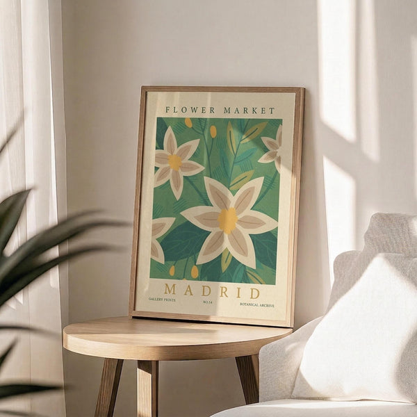 May include: Botanical poster with white flowers and green foliage displayed in thin wooden frame on round wooden side table next to beige upholstered chair. Soft natural lighting creates window shadows on neutral wall. Plant leaves visible in corner. Vintage-style artwork features cream background with decorative text above and below floral illustration.