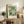 May include: Framed botanical poster with white flowers on green background displayed on rustic wooden console table alongside fern plant, books, ceramic vase and table lamp in neutral-toned living room setting with beige armchair