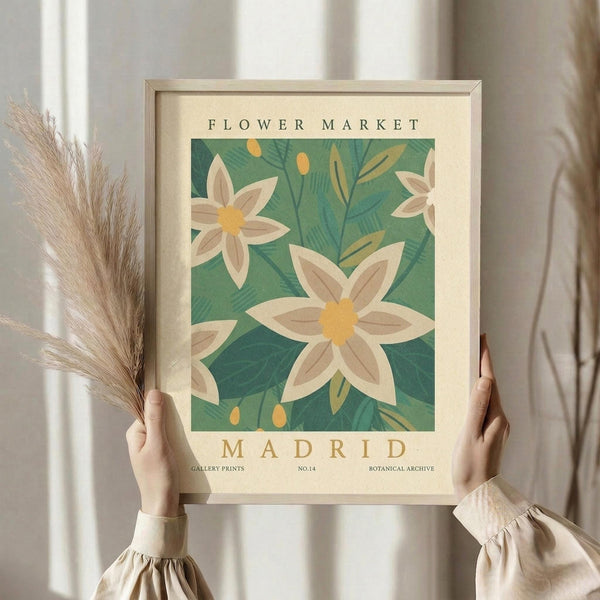 May include: Person in cream blouse holding framed botanical art print featuring white flowers with yellow centers on green leafy background. Dried pampas grass visible to left. Print has cream matting in light wood frame. Soft natural lighting against neutral curtained backdrop.