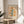 May include: Framed botanical poster with orange and yellow flowers on cream background, displayed on round wooden side table next to beige upholstered chair. Natural lighting creates soft shadows across white walls. Green plant leaves visible in corner, creating warm minimalist home decor scene.
