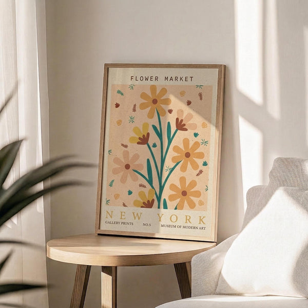 May include: Framed botanical poster with orange and yellow flowers on cream background, displayed on round wooden side table next to beige upholstered chair. Natural lighting creates soft shadows across white walls. Green plant leaves visible in corner, creating warm minimalist home decor scene.