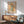 May include: Framed botanical poster with warm beige background featuring stylized flowers in yellow, orange and brown tones with green stems, displayed above a mid-century modern wooden credenza next to a sage green upholstered armchair with wooden frame in a bright living room with white walls and natural sunlight streaming across the scene.