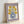 May include: Framed vintage-style botanical poster leaning against white wall showing yellow flowers with burgundy centers in blue spotted vase on cream background with purple border, displayed in natural wood frame with visible texture and grain